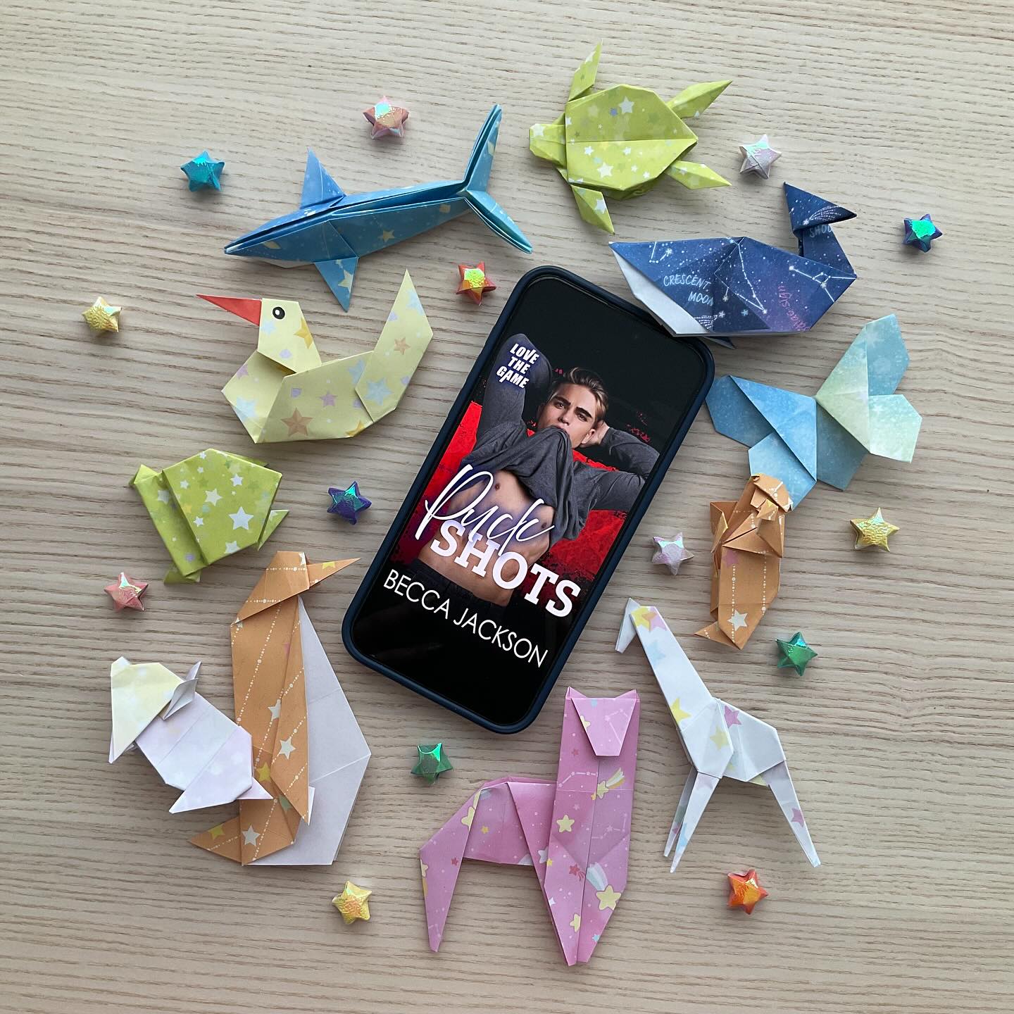 The model book cover for 'Puck Shots' by Becca Jackson displayed on an Apple iPhone 15 Pro Max, which lays scattered amongst origami animals and stars, on a wooden table backdrop. The model on the book cover has his arms above his head and the bottom of his shirt caught in his teeth in an alluring manner.