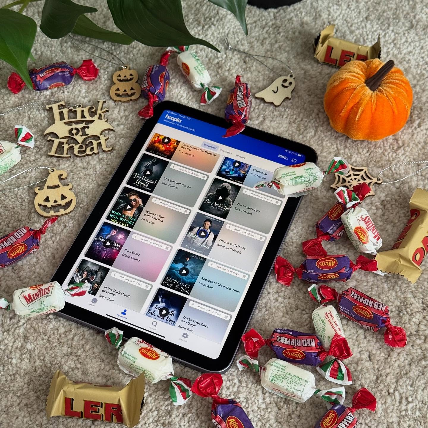 An iPad lays on a beige carpet open to the hoopla app, showing a number of borrows. The iPad is surrounded by an array of Halloween themed decorations and a scattering of sweets. Some leaves curl in from the top edge.